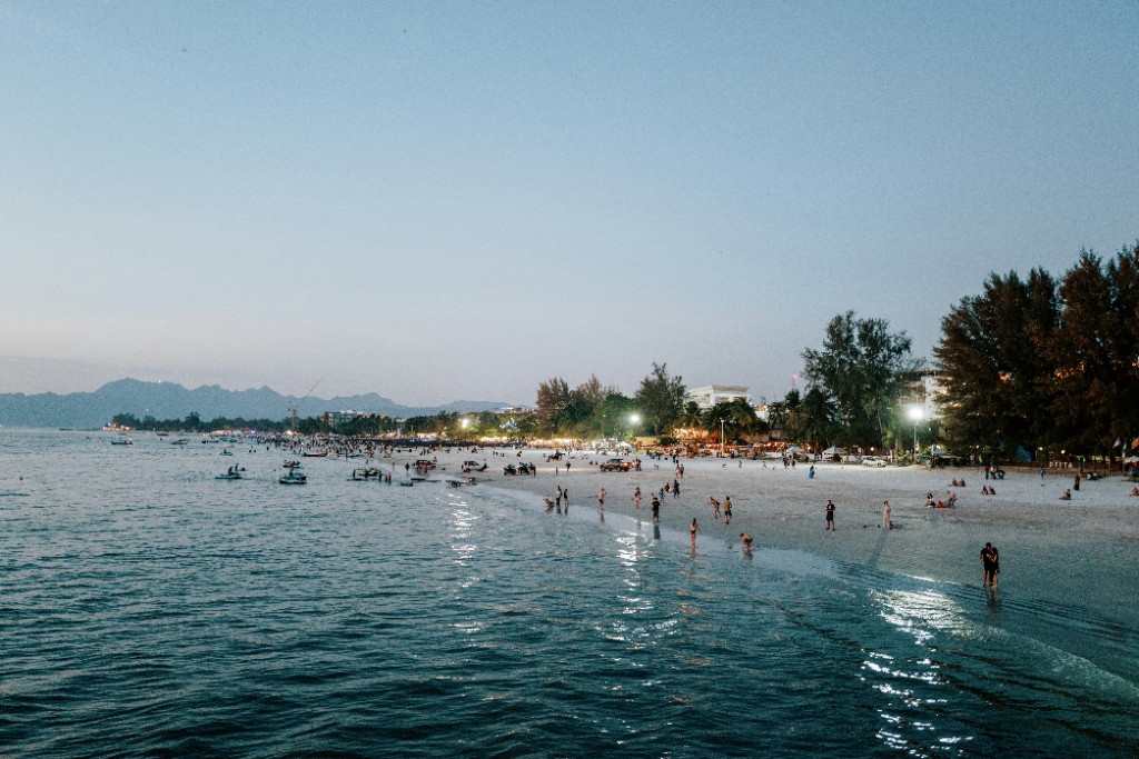Wide Langkawi shoreline at dusk with people on the sand and shore lights reflecting on the sea
